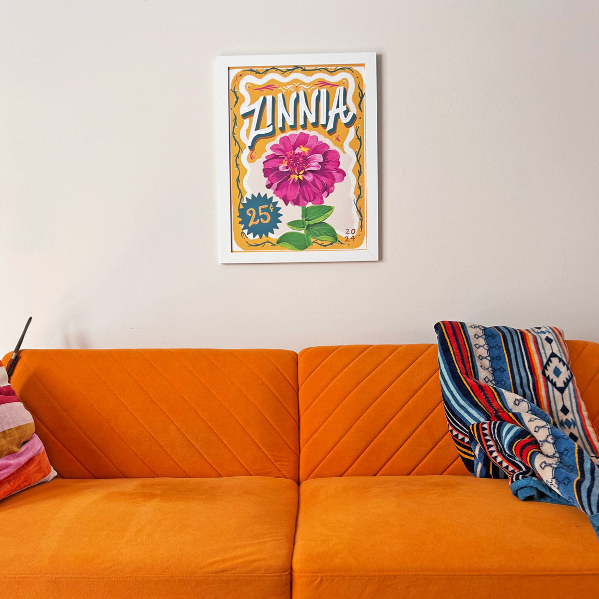 framed artwork of pink zinnia flower on the wall above a  orange sofa with a colorful blanket and pillow