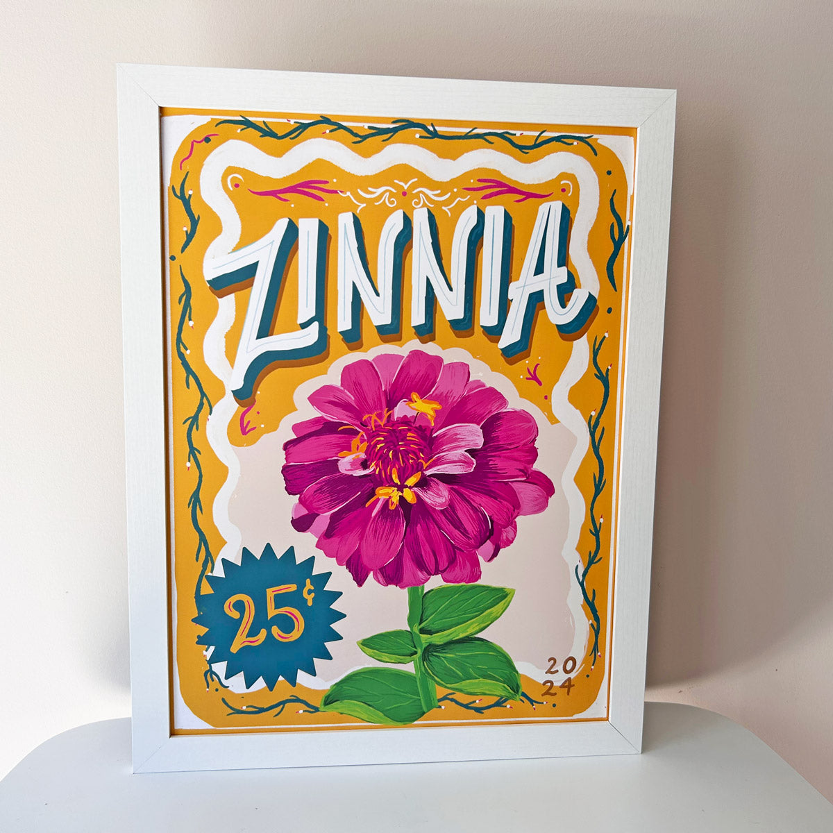 Framed artwork with a pink zinnia flower and decorative text on a white table